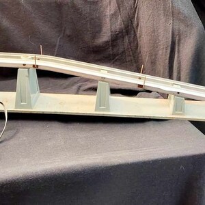 May include: A vintage toy race track set with a metal ramp and a control box with red buttons. The track is made of metal and has a gray finish. The control box is black with red buttons and a cord.