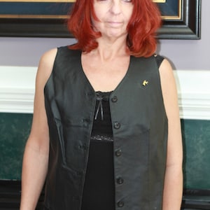 May include: A woman with red hair is wearing a black leather vest with a button closure. The vest has a small gold pin on the left side. She is wearing a black top underneath the vest.