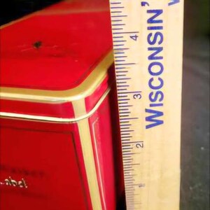 May include: A red rectangular tin box with gold trim and a white label. The label has the word "abcd" printed on it. The box is 5 inches tall.