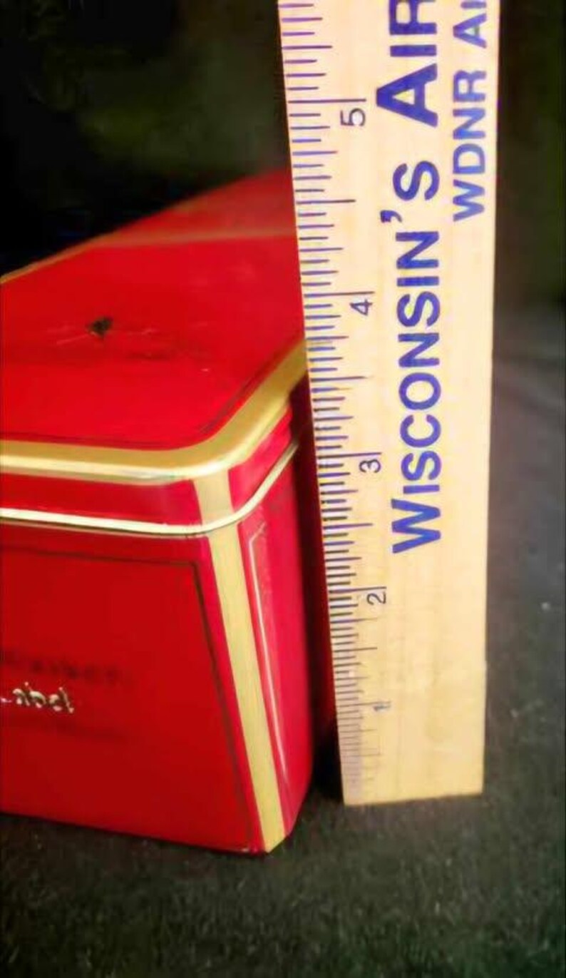 May include: A red rectangular tin box with gold trim and a white label. The label has the word "abcd" printed on it. The box is 5 inches tall.