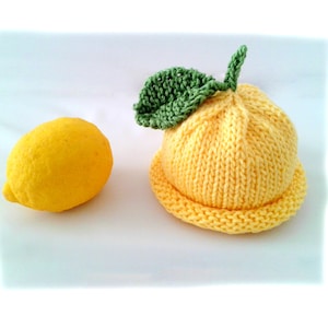 May include: A knitted yellow baby hat shaped like a lemon, complete with a green leaf and stem. A real lemon sits beside the hat. The hat is a bright yellow color, and the leaf is a vibrant green.