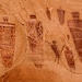 May include: A rock wall with red ochre petroglyphs depicting human figures and geometric shapes. The figures are stylized and appear to be wearing headdresses or hats. The petroglyphs are a form of ancient rock art.