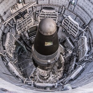 May include: A black and silver missile is positioned inside a silo, with the top of the missile visible. The silo is made of concrete and has a circular opening at the top. The missile is surrounded by metal supports and cables.