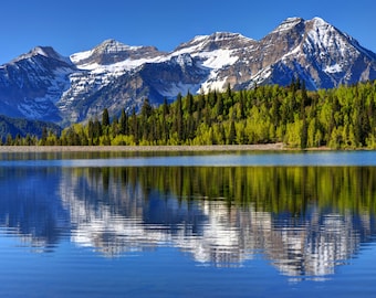 May include: A scenic view of a mountain range with snow-capped peaks reflected in a still lake. The mountains are covered in lush green trees and the lake is a vibrant blue.