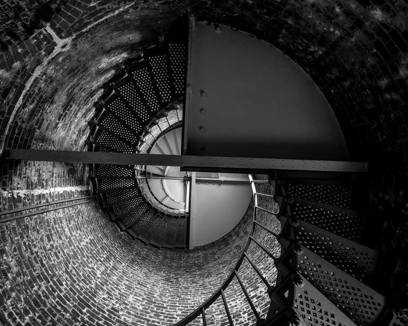 May include: A black and white photo of a spiral staircase inside a brick tower. The staircase is made of metal and has a circular pattern. The brick wall is textured and rough.