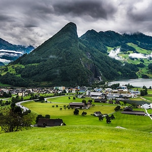 May include: A scenic view of a small village nestled in a valley surrounded by lush green mountains and a lake. The sky is overcast with clouds.