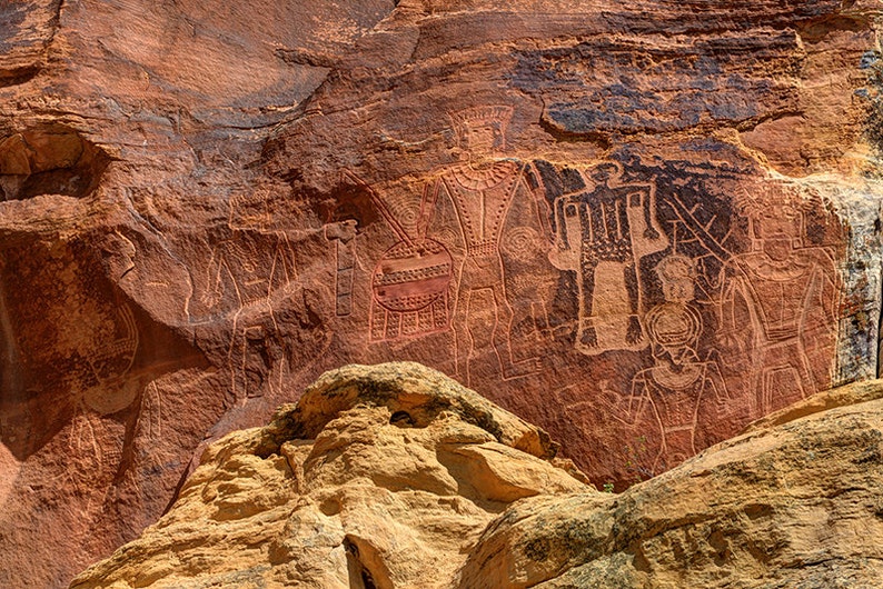 K&ouml;nnte beinhalten: Nahaufnahme einer roten Felswand mit alten Petroglyphen, die in die Oberfl&auml;che gemei&szlig;elt sind. Die Petroglyphen zeigen Figuren von Menschen und Tieren, darunter ein Kamel.