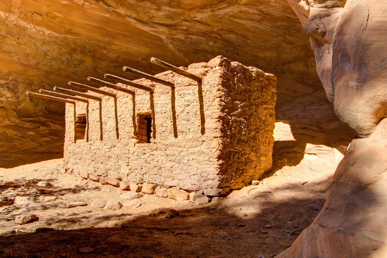May include: A stone building with a wooden roof structure, built into a sandstone cliff. The building has several windows and is made of rough-hewn stone.