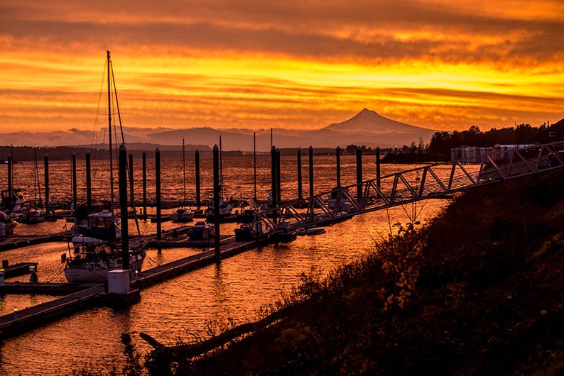 May include: A scenic view of a marina at sunset with boats docked and a mountain range in the distance. The sky is a vibrant orange and yellow, creating a beautiful and peaceful scene.