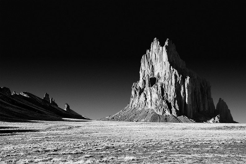 May include: A black and white photograph of a tall, jagged rock formation rising from a barren desert landscape. The rock is illuminated by the sun, casting long shadows across the ground.