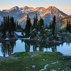 May include: A mountain lake reflecting the surrounding snow-capped peaks and evergreen trees. The water is still and clear, reflecting the sky and the mountains.