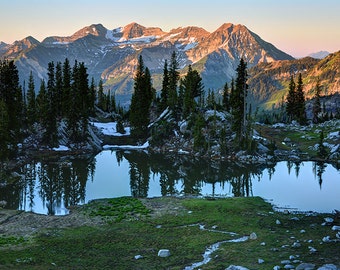 May include: A mountain lake reflecting the surrounding snow-capped peaks and evergreen trees. The water is still and clear, reflecting the sky and the mountains.