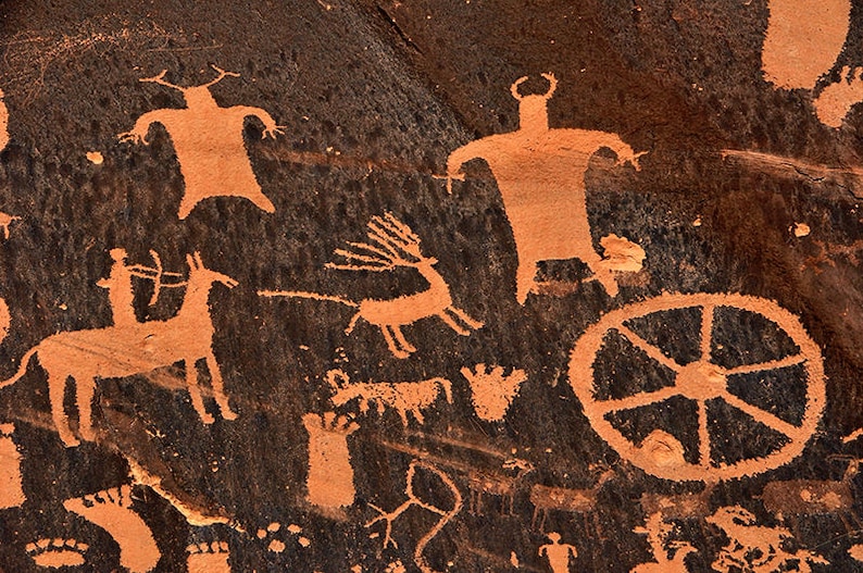 Puede incluir: Un primer plano de una pared de roca con petroglifos naranjas que representan varias figuras, incluyendo un ciervo, una persona montando un caballo y una rueda. Los petroglifos est&aacute;n tallados en la roca marr&oacute;n oscura.