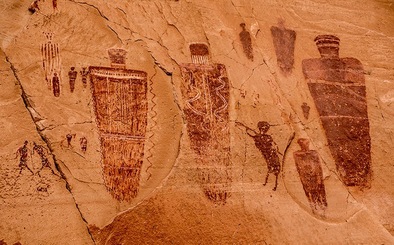 May include: A rock wall with red ochre petroglyphs depicting human figures and geometric shapes. The figures are stylized and appear to be wearing headdresses or hats. The petroglyphs are a form of ancient rock art.