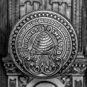 May include: A black and white photo of a metal door knob with the words "Holiness to the Lord" inscribed around a beehive design.