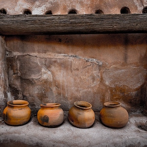 May include: Four terracotta pots sit on a ledge in front of a rustic stone wall. The pots are all different sizes and shapes, and they have a worn, aged look.