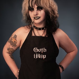 Op de afbeelding: Een zwarte schort met de tekst "Goth iHop" in wit met een pentagram symbool. De persoon die de schort draagt heeft zwart en wit haar, donkere make-up en meerdere piercings.