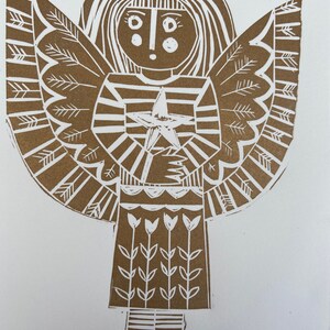 May include: A brown linocut print of an angel with a crown, wings, and a star. The angel is wearing a striped dress and has a simple face. The print is signed 'crest' in the bottom right corner.
