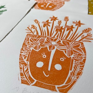 May include: A hand-carved orange print of a smiling face with a crown of flowers. The print is on white paper and says "A bunch of Daffodils" at the bottom.