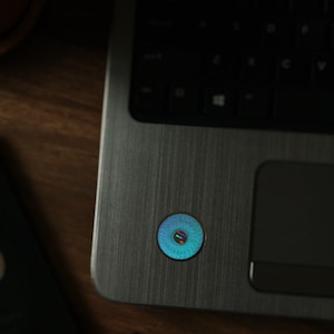 Puede incluir: Un ordenador portátil plateado con un teclado negro y una pegatina circular azul y verde en el touchpad.