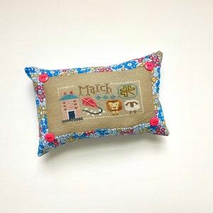 May include: A small, rectangular pillow with a floral print fabric cover. The pillow has a beige linen front with an embroidered design featuring a house, a kite, a lion, a sheep, and the word "March". The pillow is accented with four pink buttons.