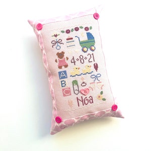 May include: Pink pillow with a white embroidered design featuring a baby carriage, toys, and the date 4-8-21. The pillow has pink buttons and the name "Noa" is embroidered at the bottom.