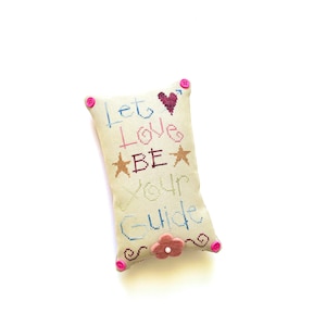 Könnte beinhalten: Ein rechteckiges, cremefarbenes, kreuzgesticktes Kissen mit den Worten "Let Love Be Your Guide" in verschiedenen Farben. Das Kissen hat ein Herz- und Stern-Design, rosa Knöpfe an den Ecken und eine Blumenverzierung.
