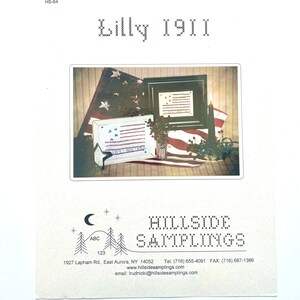 May include: A vintage-style cross-stitch pattern featuring a red, white, and blue American flag with the text "Lilly 1911". The pattern is framed and displayed on a table with other patriotic decorations.