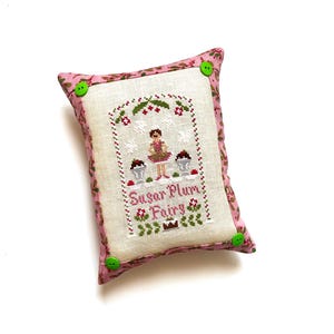 May include: A rectangular cushion with a pink floral border and green buttons. The centre features a cross-stitched design with a fairy, cupcakes, and the words "Sugar Plum Fairy."