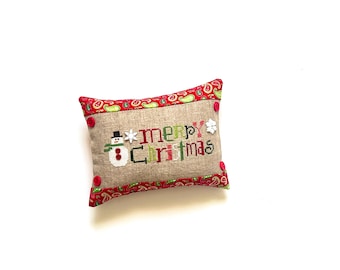 May include: A rectangular Christmas pillow with a beige linen-like fabric. The pillow features the words "Merry Christmas" embroidered in green, red, and white, along with a snowman and snowflake design. The edges are trimmed with a red paisley pattern.