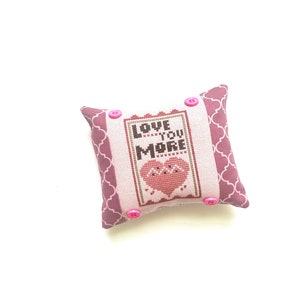 Könnte beinhalten: Ein kleines rosa und weißes Kissen mit einem Kreuzstich-Design, auf dem "Love You More" mit einem rosa Herz in der Mitte steht. Das Kissen hat rosa Knöpfe an den Ecken.