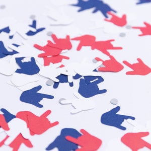 May include: Red, white, and blue hand-shaped confetti scattered on a white surface. The confetti is in the shape of the "I love you" hand sign, with small silver confetti pieces scattered throughout.