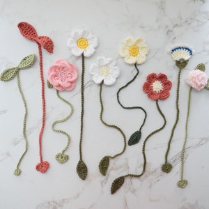 Puede incluir: Un conjunto de nueve marcapáginas de flores de ganchillo en varios colores, incluyendo rosa, blanco y crema. Las flores están unidas a tallos largos y verdes con hojas.