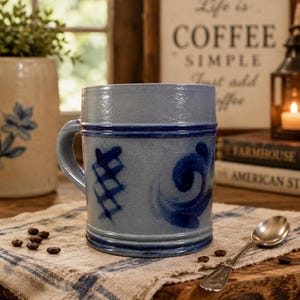 May include: A cylindrical light blue ceramic mug featuring a dark blue horizontal band near the top and bottom, decorated with hand-painted dark blue cross-hatch and swirl patterns. The mug has a sturdy side handle and a smooth, glazed finish.