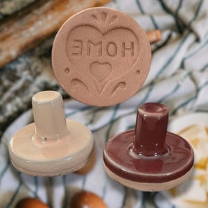 May include: Two ceramic cookie stamps, one with a pink glaze and the other with a dark red glaze. The pink stamp has a heart design with the word "HOME" inside. The red stamp has a smooth, rounded top.