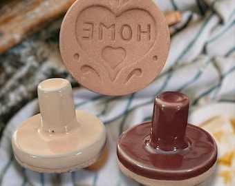 May include: Two ceramic cookie stamps, one with a pink glaze and the other with a dark red glaze. The pink stamp has a heart design with the word "HOME" inside. The red stamp has a smooth, rounded top.