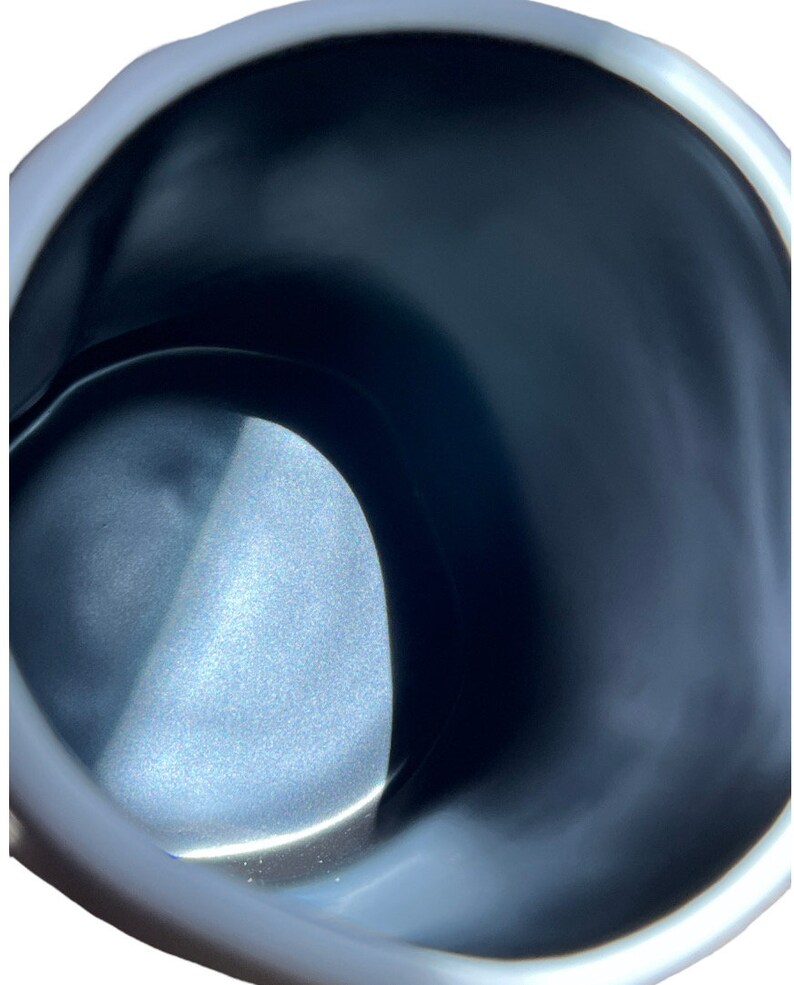 May include: Close-up of a white and black ceramic mug. The interior is dark, with a glossy finish. The rim of the mug is white and has a slightly irregular shape. The mug appears to be empty.