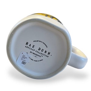 May include: White ceramic mug with a handle, featuring the text "RAE DUNN ARTISAN COLLECTION BY MAGENTA". The mug has a simple design and a small sticker with product information.