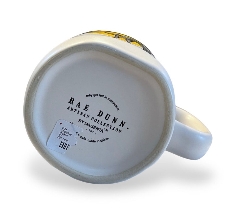 May include: White ceramic mug with a handle, featuring the text "RAE DUNN ARTISAN COLLECTION BY MAGENTA". The mug has a simple design and a small sticker with product information.