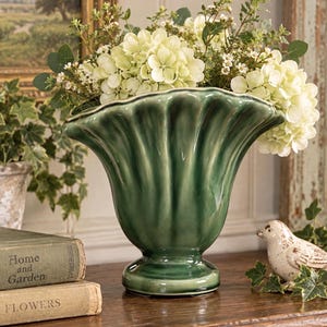 May include: A glossy, dark green ceramic vase with a wide, scalloped, fluted rim and a pedestal base. The vase is filled with a large arrangement of cream-colored hydrangea flowers and green foliage, creating a full, rounded display.