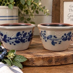 May include: Two matching beige stoneware bowls featuring a hand-painted blue berry and leaf vine pattern. Each bowl has a stamped maker's mark on the front and a smooth, rounded rim. The interior of each bowl is finished in a dark, solid black glaze.