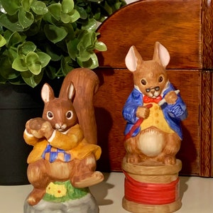 May include: Two ceramic figurines of squirrels. One squirrel is holding a nut and wearing a yellow jacket with a blue and white plaid shirt. The other squirrel is wearing a blue jacket with a yellow vest and a red bow tie. The squirrel is holding a paintbrush and wearing glasses.
