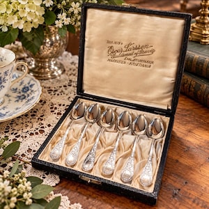 May include: A set of six silver spoons with ornate floral handles arranged in a black presentation box. The interior lid features gold text reading TELE 1878 Oscar Larssen Guldsmed og Iravir EXALIE URETAFUR MARKVEIEN-KRISTIANIA.