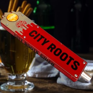 May include: A red and tan tap handle with the words "CITY ROOTS" in large black letters. The handle has a cityscape design and the words "PUMPKIN CIDER" in a circle. The tap handle is in front of a glass of beer.