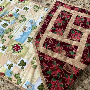 May include: Two quilted table runners with a Christmas theme. One runner features a red background with poinsettia flowers and gold accents. The other runner has a beige background with a winter scene and green ivy.