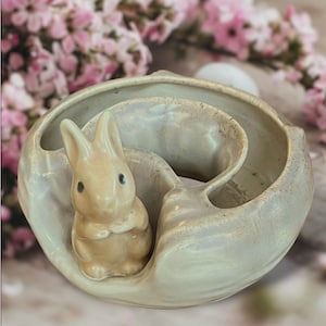 May include: A ceramic Easter bunny figurine inside a cream-colored, shell-shaped dish. The bunny is light brown with black eyes and is positioned in the center of the dish. The dish has a textured surface and a curved design, perfect for Easter decor.