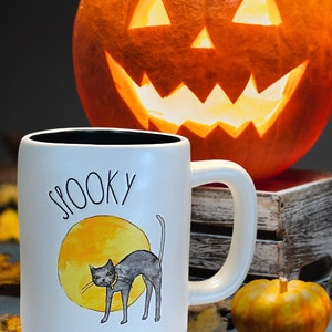 May include: White ceramic mug with the word "SPOOKY" and a black cat illustration in front of a yellow moon. A carved orange pumpkin with a lit face is in the background, along with a small pumpkin and autumn leaves.