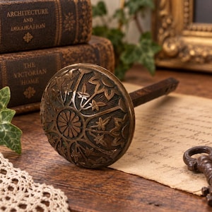 May include: A round, antique bronze door knob featuring a detailed, embossed floral and leaf pattern radiating from a central circular motif. The knob includes a square-shaped metal spindle extending from the back, showcasing a dark, weathered finish.