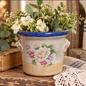 May include: A ceramic planter with a blue rim and two side handles, featuring a floral decal of white, pink, and purple flowers on a beige body. The planter is filled with a cluster of green foliage and small white flowers.