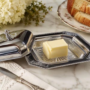 May include: A silver-toned metal butter dish with a removable lid and a clear glass insert. The base features an ornate, textured border, while the lid has a small finial handle. A single square block of yellow butter sits inside the dish.